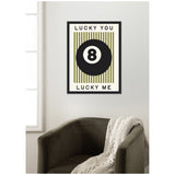 Framed print of a black 8 ball centered on vertical olive green stripes, bordered by 'LUCKY YOU' above and 'LUCKY ME' below in black text, hanging above a dark gray armchair with a cream knit throw.