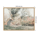 Framed canvas showing a cheetah lying on grass with blurred trees and foliage in soft, muted natural tones as background.