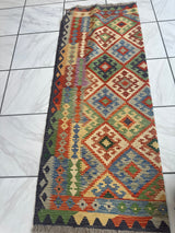 Rectangular handwoven Afghani wool kilim rug featuring multicolored geometric diamond patterns and vertical stripes in rich earthy tones on white tiled floor.