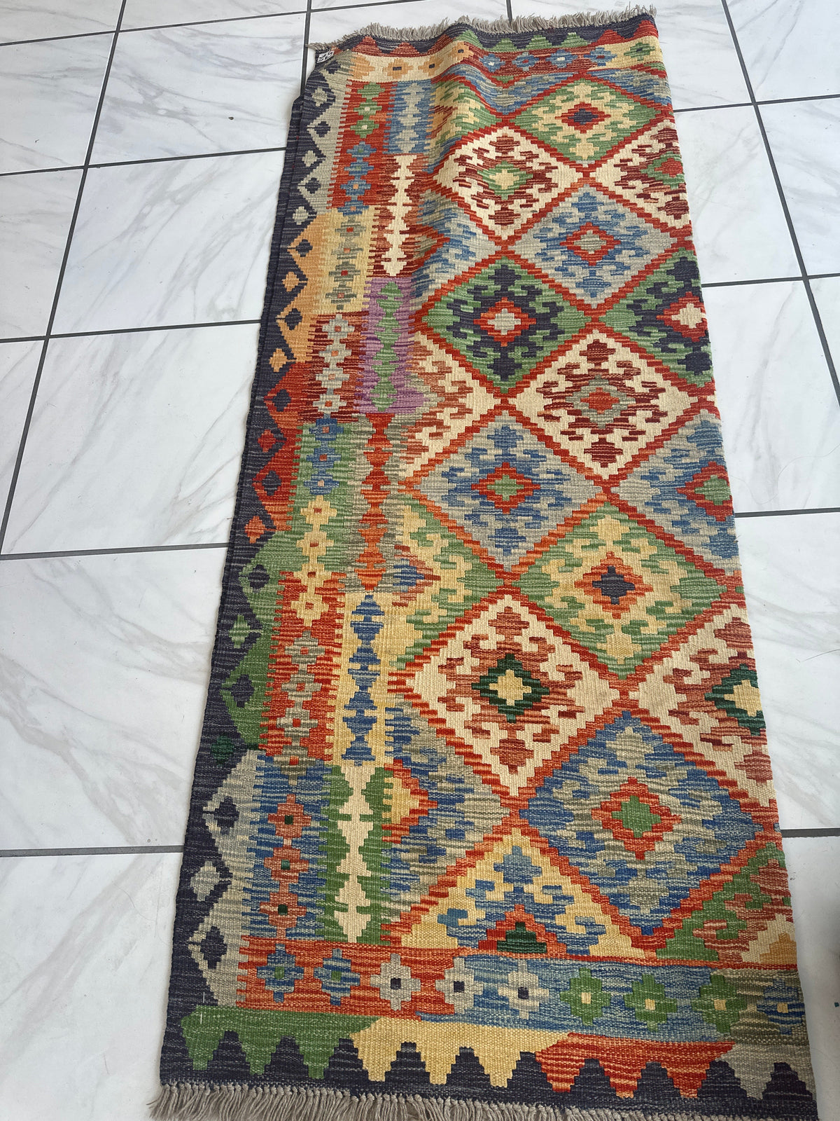 Rectangular handwoven Afghani wool kilim rug featuring multicolored geometric diamond patterns and vertical stripes in rich earthy tones on white tiled floor.