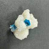Clusters of bright blue cavansite balls nestled within translucent white stilbite crystals on a gray background, showing intricate, sparkling formations.