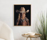 Framed portrait of a brown horse wearing a tan western cowboy hat and saddle against a black background, hung on a white wall above a round brass table and chair.