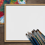 Square blank canvas framed in light wood placed diagonally on a wooden surface with a colorful paint palette at the top left and a cluster of varied paintbrushes at the bottom right.