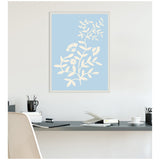 Framed canvas print of pale blue background with cream botanical floral design featuring leaves and flowers, hanging above a minimalist desk with office supplies.