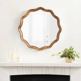 Round scalloped mirror with a natural wood frame featuring gentle wavy curves, hung above a white mantel decorated with a candle and potted plant.