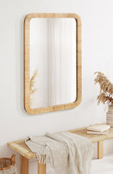 Rectangular wall mirror with rounded corners and a light wood frame, hanging above a woven bench topped with a beige throw blanket and books, next to a vase of dried pampas grass.