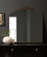 Large traditional arch-shaped mirror with ornate bronze top detail, mounted on a dark wall above a black table holding a textured white vase and candle.