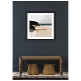 Abstract coastal landscape with beige sand, dark rocky slope on left, and soft blue-gray sky, framed and hung on a dark wall above a wooden bench with two woven baskets underneath.