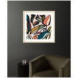 Framed abstract botanical wall art with flowing shapes in teal, pink, yellow, and coral tones on a pale blush background, hung above an olive green armchair and black side table.