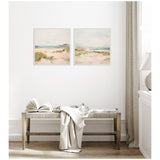Two square pastel landscape paintings with soft sandy dunes, pale greenery, and distant mountains framed on a white wall above a wooden bench with a draped blanket and open book.