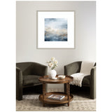 Square abstract wall art with blue, grey, and beige tones above a sitting area with two dark green armchairs and a round wooden table with white vase and books.