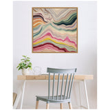 Abstract landscape artwork with layered waves in cream, pink, green, yellow, and orange tones framed and hanging above a minimalist wooden desk with a plant and gray chair.