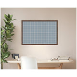 Wall-mounted rectangular pinboard with light blue plaid fabric framed in dark wood above a white desk with stacked books and a potted plant to the left