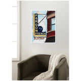 Framed canvas wall art featuring a stylized street sign reading 'Pardon my French' with a brick building and sky background, displayed above a dark gray armchair with a white knit throw.