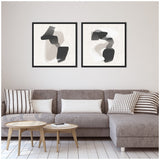 Two framed abstract canvas prints with overlapping gray and black shapes in soft beige backgrounds, displayed above a gray sectional sofa with patterned pillows.