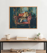 Painting of a lion reclining on a red armchair against a dark, abstract jewel-toned background, framed in light wood and hung above a wooden table with neutral decor.