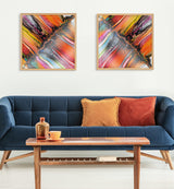 Two framed abstract prints with vibrant diagonal streaks of orange, pink, yellow, black, and white hanging above a blue cushioned sofa with mustard and rust pillows, beside a wooden coffee table with decorative bottle and cup.
