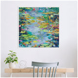 Abstract painting of water lilies in vibrant blues, greens, and hints of pink, framed and hung above a wooden desk with a gray chair and potted green plant.