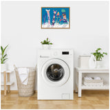 Framed artwork above a white front-loading washing machine shows colorful skis and snowboards standing in snow against a bright blue sky, adding vibrant contrast to a minimalist laundry space.