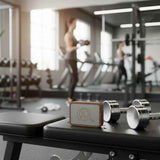 Small wooden and metal sensor device with a logo on a black gym bench next to silver dumbbells; blurred person exercising in background.