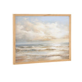 Framed print depicting a beach scene with gentle waves under a cloudy sky at sunset, featuring soft beige, blue, and white tones in a natural wood frame.