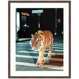 A tiger walking across a city crosswalk at night with dark buildings and illuminated signs in the background, framed with a wooden border.