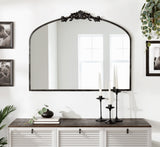 Arched wall mirror with dark metal frame featuring a central ornamental design, hanging above a white cabinet with dark wooden top, styled with candles and plants.