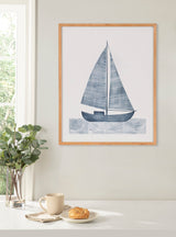 Minimalist framed print of a navy blue sailboat with textured sails above stylized water waves, hanging on a light beige wall next to a window and a table with a plant, cup, and pastry.