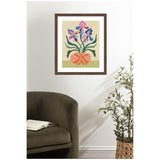 Framed abstract art of purple and pink iris flowers in a round orange patterned vase, hanging on a white wall above a dark green armchair with an open book and a potted olive tree nearby.