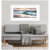 Horizontal painting of teal-green ocean waves crashing under a cloudy sunset sky, framed in white and hung on a beige wall above a gray sofa with a soft gray throw and cushions.
