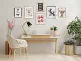 Modern workspace with a light wood desk holding a black lamp, clock, and small plants, beige upholstered chair, and six framed prints including bows and animal illustrations on the wall above.