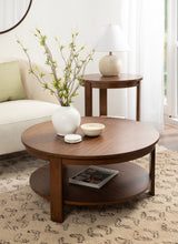 Round wooden coffee table with lower shelf holding a magazine, topped with a white vase of green branches and two small bowls, beside a round wood side table with ceramic lamp.