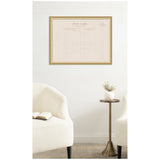 Framed pinboard resembling a vintage linen postcard with beige fabric and muted text, hanging on a white wall above minimalist cream armchairs and a small brass side table with a white vase.