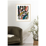 Abstract botanical art print with overlapping colorful ficus leaves in teal, mustard, coral, and black outlines on a pale pink background, framed in light wood and hung above a green chair.