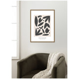 Framed abstract wall art with black leaf-like shapes on a beige background, titled 'Les Fleurs,' hung above a dark green armchair with a textured cream throw.
