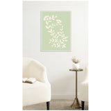 Framed wall art with cream floral silhouette on pale green background above a small round brass table and two white upholstered chairs, one with open book.