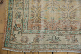 Corner of a 5.5x9 vintage distressed Oushak carpet with faded earth tones, muted turquoise accents, and intricate floral border patterns on wooden flooring.