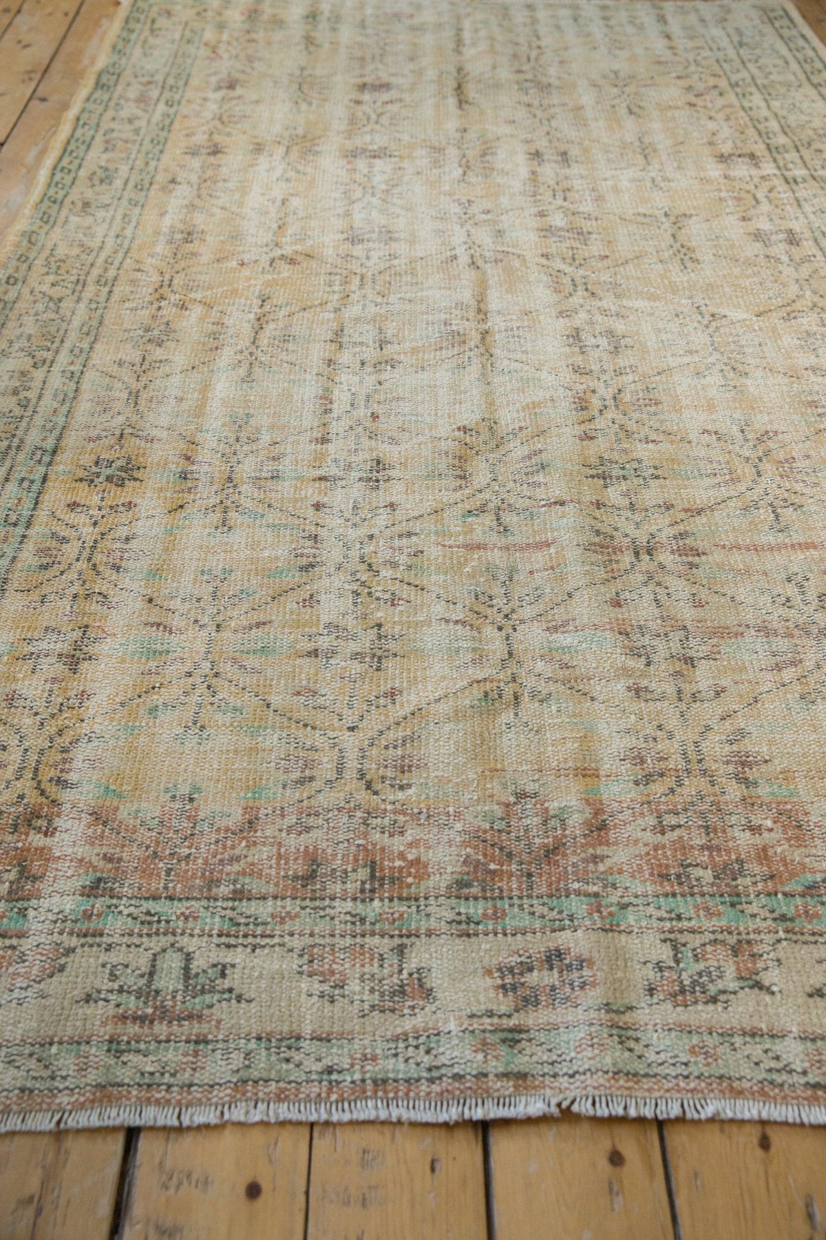 Vintage distressed Oushak carpet in faded beige and muted teal with floral motifs and border, laid on a wooden floor.