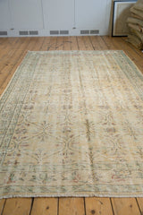 Vintage distressed Oushak carpet with faded beige background and soft green floral border, laid on light hardwood floor beside stacked rugs and framed artwork.