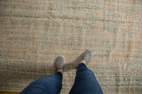 Worn vintage Oushak carpet in muted beige and green tones with faded traditional patterns, viewed from above by a person wearing gray slip-on shoes and blue jeans.
