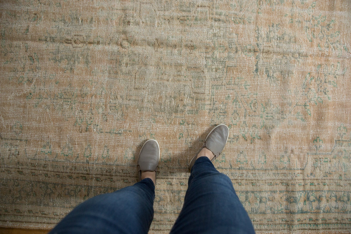 Worn vintage Oushak carpet in muted beige and green tones with faded traditional patterns, viewed from above by a person wearing gray slip-on shoes and blue jeans.