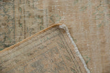 Close-up of a vintage distressed Oushak carpet in muted tan and sage green tones with faded geometric patterns and a frayed edge corner folded over.