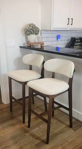 Two mid-century style bar stools with dark wood legs and curved backs upholstered in cream textured fabric, placed by a kitchen counter with black countertop and white subway tile backsplash.