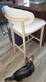 Light wood mid-century modern bar stool with white upholstered cushioned seat and backrest, shown on wood floor near a dark wood counter, small dog beneath stool.