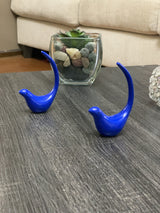 Two small blue bird figurines with elongated upward tails on a dark wood table, accompanied by a glass planter with succulents and pebbles in the background.