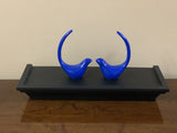 Two small, glossy blue bird figurines with long, curved tails facing each other on a black rectangular tray, displayed on a wooden surface against a beige background.