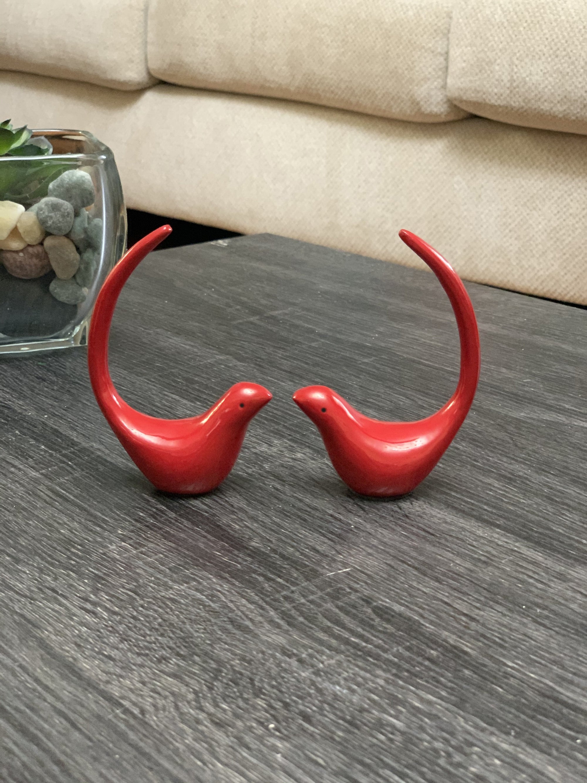 Two red metal bird figurines positioned on a light-colored surface, one with a ring on its tail