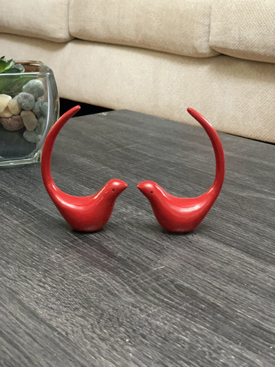 Two red metal bird figurines positioned on a light-colored surface, one with a ring on its tail