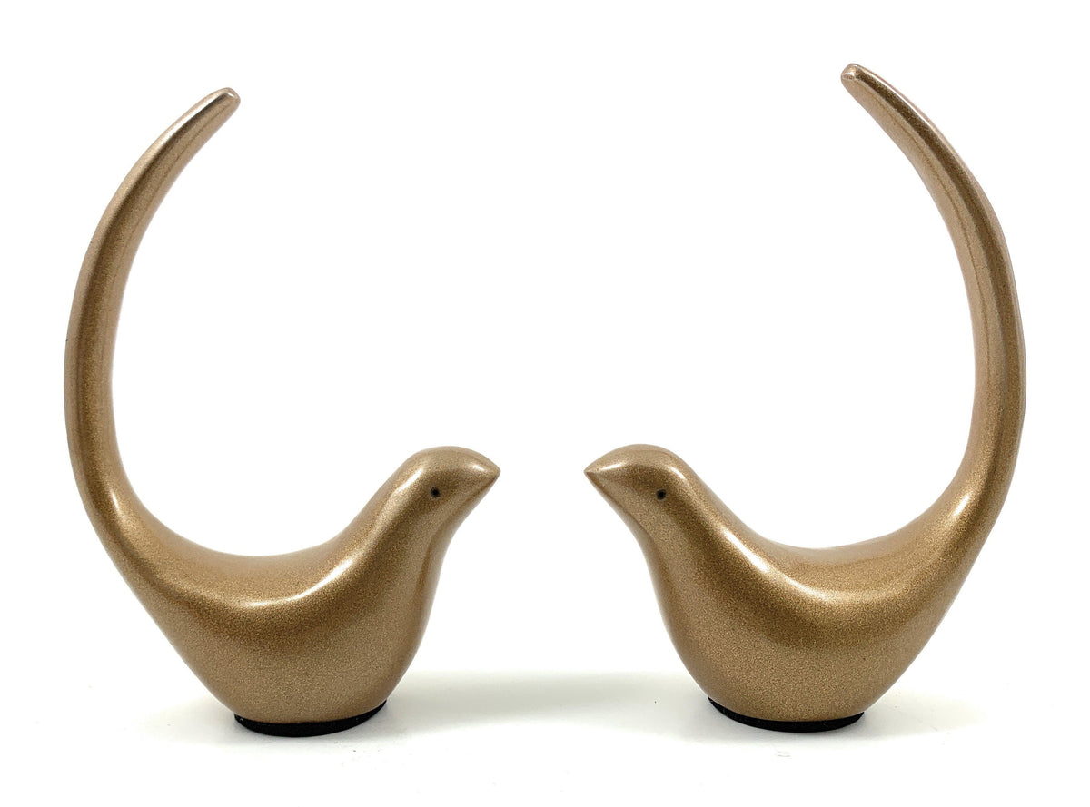 Pair of smooth, gold-toned bird figurines with long, curved tails arching upwards, facing each other against a white background.