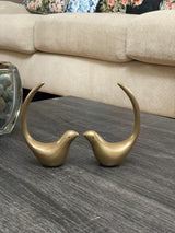 Two small golden bird figurines facing each other with long curved tails, placed on a dark wood table in front of a beige sofa with floral cushions.